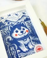 Linogravure bleue d’un chien Akita assis dans un champ, vu de trois quarts, avec des montagnes japonaises et des libellules en arrière-plan