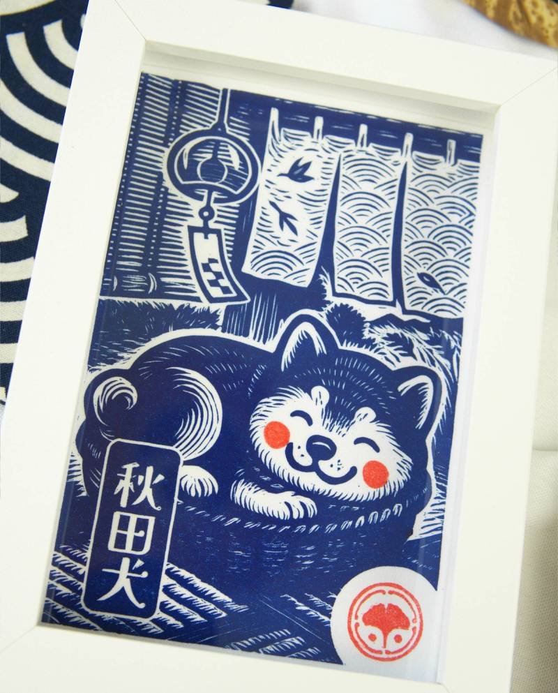 Linogravure bleue d’un chien Akita endormi dans un panier sur un tatami, avec un noren flottant et un furin suspendu en arrière-plan