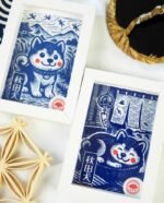 Linogravure bleue d’un chien Akita endormi dans un panier sur un tatami, avec un noren flottant et un furin suspendu en arrière-plan